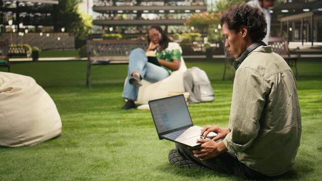 The international student uses a laptop with coding content on a campus bench while classmates study in the background for outdoor learning, for coding and automation study in an academic environment.