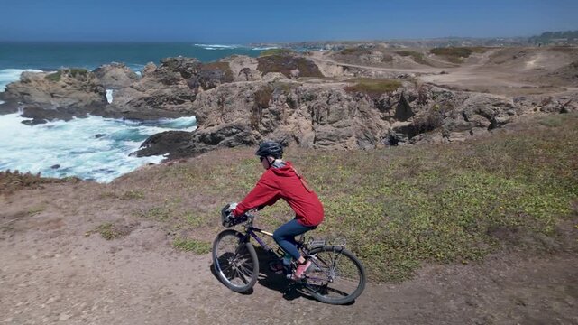 A woman enjoys biking along the cliffs in Fort Bragg, California, with waves crashing against the rocks below.