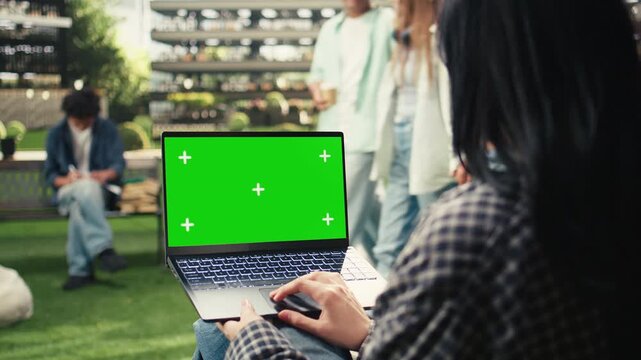 Green screen phone mockup is held close to the camera in an outdoor campus setting, with a blurred student in the background and copy space for mobile screen replacement and digital education content.