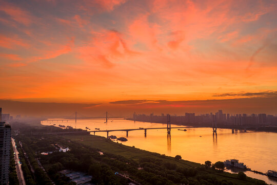 Spectacular aerial dawn over the Yangtze River in Wuhan, China. Dramatic fiery clouds reflect on golden waters, highlighting the modern city skyline, lush green riverbanks, and cable-stayed bridges.