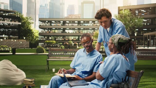Diverse med school students learn together with coursework in a group study session, with classmates gathering on campus bench, during peer tutoring.