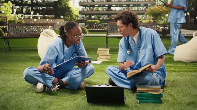 Medical students in blue scrubs discuss coursework beside a laptop and open textbooks on campus lawn, using clipboard to share notes and research ideas, during med school peer tutoring.