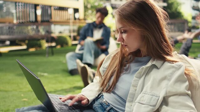 Student sits on a beanbag holding an open book and a laptop, looks up during independent outdoor study and keeps a notebook nearby on a university campus with peers in the background.