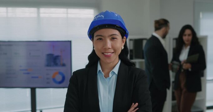 Portrait of confident Asian lady engineer in blue hardhat smiles crossing arms in boardroom. Young Korean female construction manager presents building project