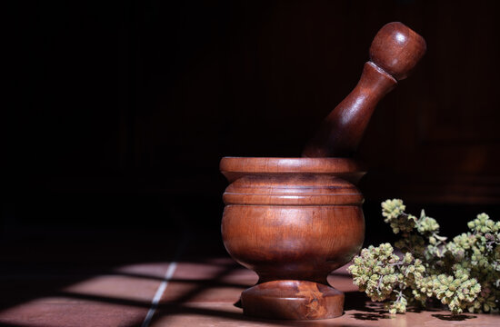 A mortar with a wooden pestle sits on a tile floor. Freshly harvested oregano lies next to it. The sun shines into the room, casting shadows.