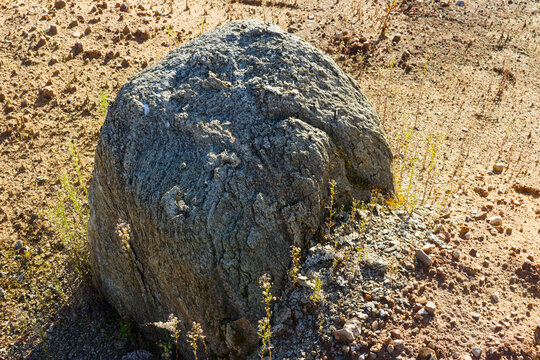 Large granite-gneiss boulder crumbling into coarse sandy grit and weathered debris, resting on the arid surface of a gravel quarry floor under bright sunlight