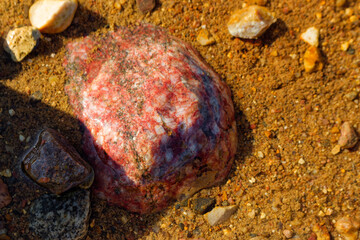 Vibrant red granite boulder rich in potassium feldspar resting in shallow water against a backdrop of sandy gravel texture in a picturesque quarry, detailed natural geological surface © PhotoChur