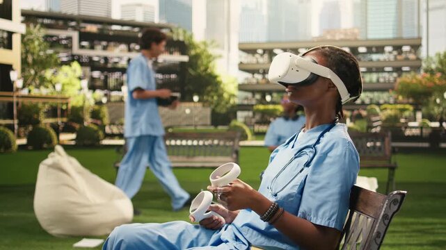 The woman medical student wears VR glasses and raises the controllers during medical training on campus, sitting on a bench in blue scrubs, using virtual reality for healthcare learning.