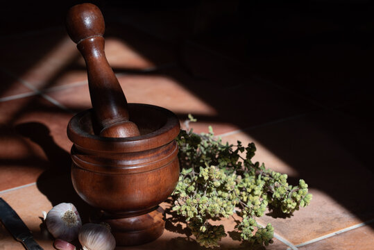 A mortar with a wooden pestle sits on a tile floor. Freshly harvested oregano and garlic lie next to it. The sun shines into the room, casting shadows.