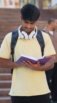 Young indian male student reading book on campus steps