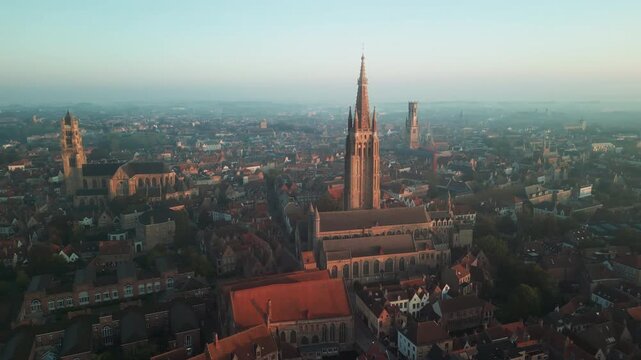 Aerial panorama of Bruges, Belgium, with the Church of Our Lady towering above medieval rooftops and landmark spires across the city in warm sunrise haze.