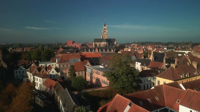 Aerial panorama of central Bruges, Belgium, shows a historic church tower above tightly packed red rooftops, leafy courtyards and medieval urban architecture.