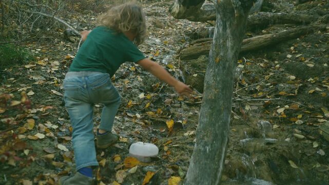 Young boy scrambling down a rocky slope toward an autumn creek holding a tree trunk
