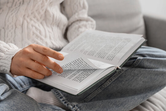 Person in a cozy sweater and jeans turning a page of an open book while reading indoors, close up, the close up shot focuses on the hand and the book