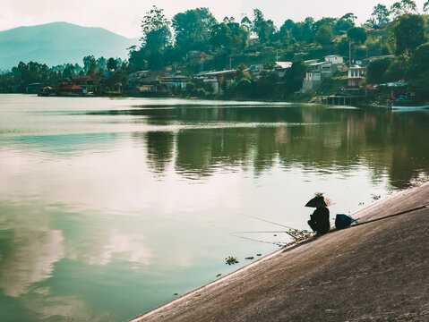 Person Fishing on Concrete Embankment by Lake with Mountain Village
