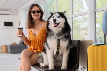 Young woman in sunglasses with a husky dog and coffee cup waiting in a bright airport lounge, a yellow suitcase and passport are visible nearby, indicating travel © Pixel-Shot