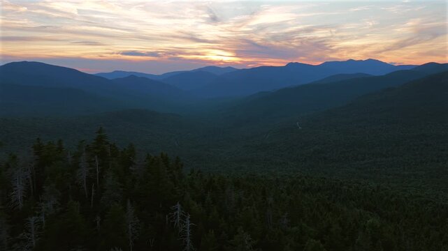 Slow aerial pan above evergreen treetops reveals a vast forested valley and layered White Mountain ridges beneath a dramatic summer sunset along New Hampshire&rsquo;s Kancamagus Highway. Text or logo room.