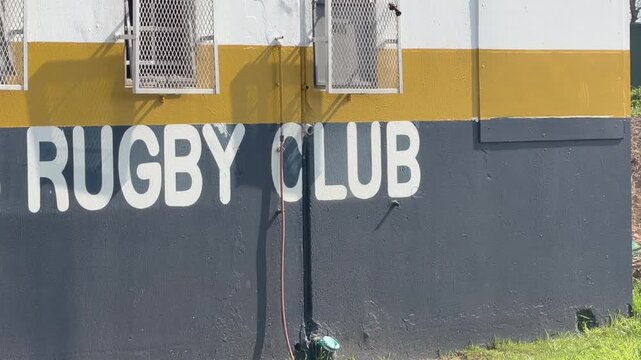 Close-up shot of a rugby club building exterior featuring bold white &ldquo;RUGBY CLUB&rdquo; lettering painted on a navy blue wall, with a mustard yellow stripe and white upper section