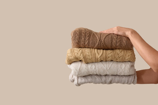 Womans hand holding a neatly folded stack of warm cable knit sweaters in neutral colors, isolated on a beige background