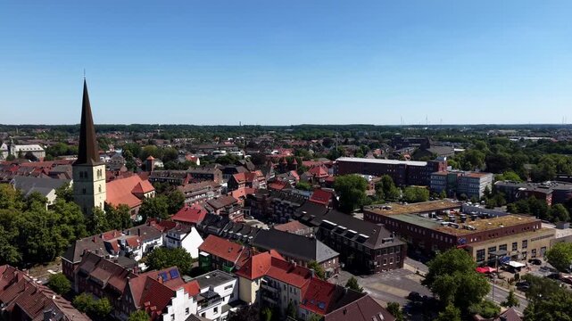 Tall church spire rises above red-roofed old town near L&uuml;dinghauser Tor in D&uuml;lmen, North Rhine-Westphalia, dense residential streets stretch toward distant tree line. Aerial drone flyover shot
