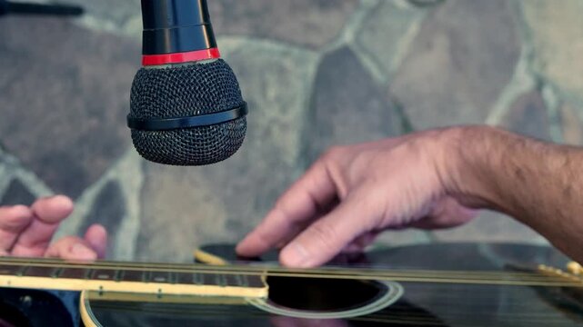 POV (zoom in): man playing acoustic guitar in front of a microphone. A guitar player in front of a microphone. A guitarist strumming a guitar. An artist plucking a guitar. A musician playing guitar
