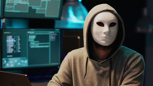 Anonymous individual wearing a white mask and hooded sweatshirt intensely working on multiple computer screens displaying code and data in a dark room
