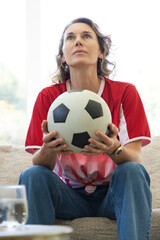 Fototapeta premium Woman wearing red-white soccer jersey sitting on sofa holding black-white ball by window watch seen