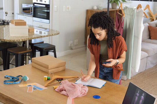 Woman wearing rust shirt standing at wood table in kitchen measuring fabric with phone, copy space