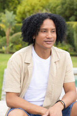 Fototapeta premium African American man sitting on wooden bench in garden, wearing beige shirt and bracelet