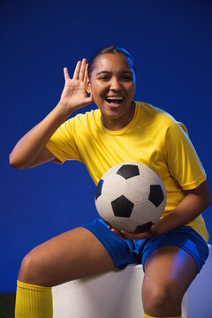 Adult African woman sitting in studio holding traditional soccer ball cupping ear in yellow jersey