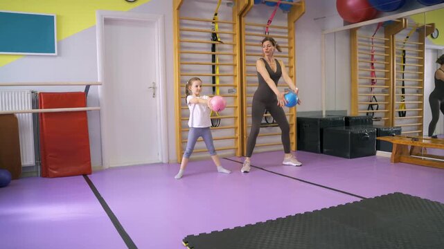 PE teacher and little girl exercising together in a gym, placing rubber ball above their head with legs spread