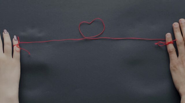 Two hands connected by a red string of fate forming a heart shape on a dark background.