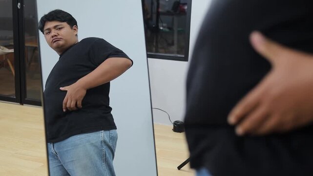 Young plus-size Asian man looking in mirror, feeling unhappy about his body image
