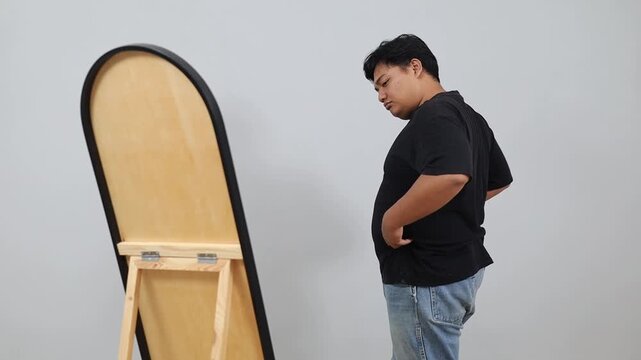 Unhappy Young plus-size Asian man looking in mirror, feeling sad about his body image