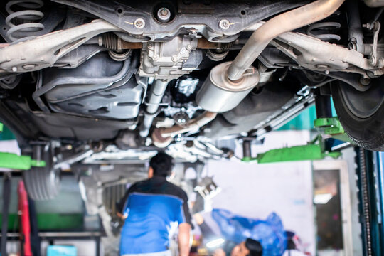 Selective focus to automotive mechanic and detailed view of a vehicle's undercarriage, rear suspension, exhaust system, and rear differential raised on a hydraulic lift in an auto repair workshop.