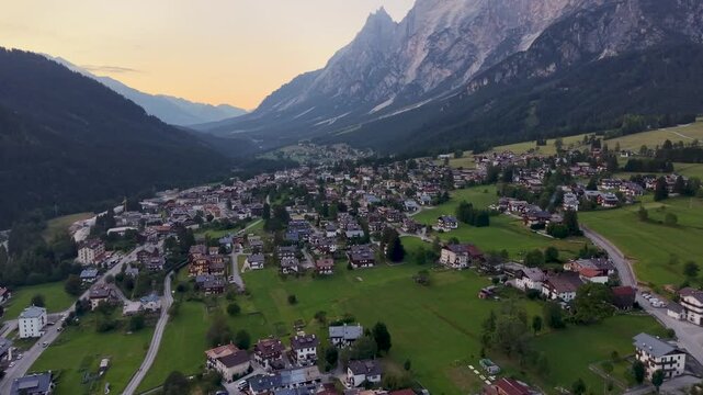 Cinematic drone flyover above the alpine village of San Vito di Cadore at sunset, surrounded by lush meadows and the dramatic peaks of the Dolomites in northern Italy