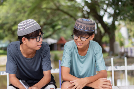 Two smiling Muslim teenage boys wearing traditional caps and talking together while sitting in a park with a blurred green background.