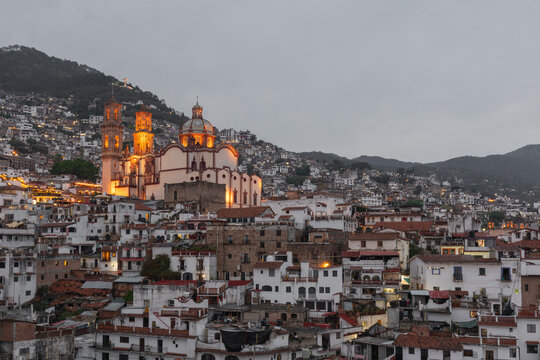 Evening cityscape of Taxco de Alarc&oacute;n with the illuminated twin towers of Santa Prisca Church rising above whitewashed colonial buildings and steep mountain slopes in Guerrero, Mexico