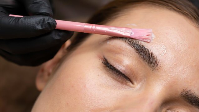 A close-up photo of a person's face during a professional eyebrow waxing or tinting treatment, with a gloved hand holding a pink brush over the eyebrow area