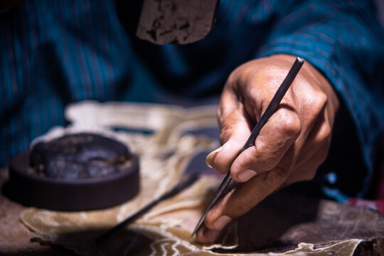 Focus on artisan hand holding thin metal chisel to carve intricate pattern on wayang kulit puppet sheet