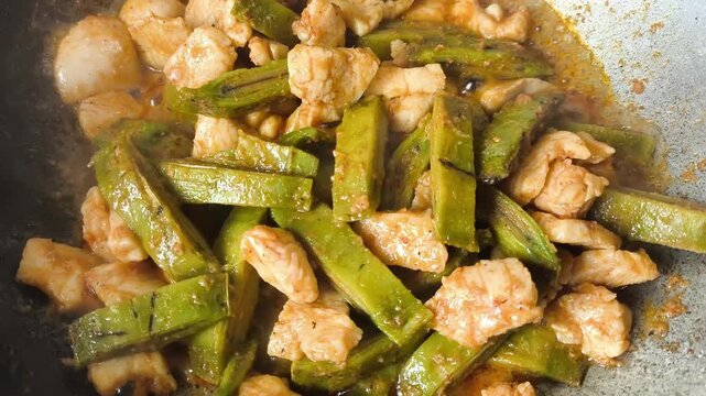  A close-up shot of cooking a unique Thai dish, stir-fried chicken breast with sliced local green fruit called Oroxylum indicum (Phayom) inside a rustic metal wok. The video captures the hot sauce bub