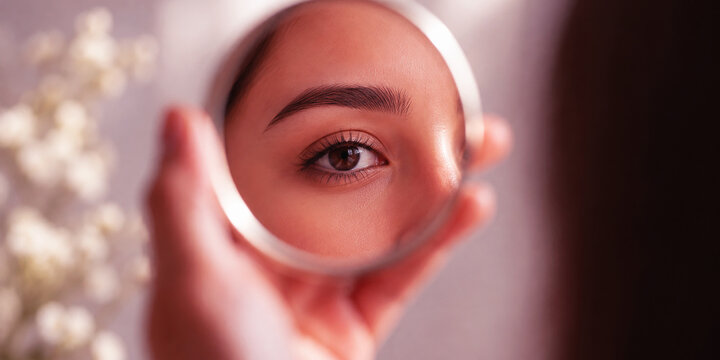 Close up of a beautiful young woman looking into a small round mirror reflecting a clear brown eye