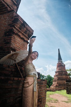 Asian teenage girl tourist dressed in traditional Thai outfit enjoying taking selfie with mobile phone at old brick ancient ruins, holiday travel in Ayutthaya historic park,  travel destination.