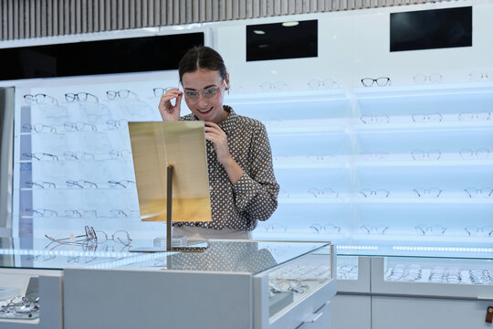 One attractive White female customer chooses and tries on eyeglasses, looks in mirror for improved eyesight for individual lifestyles, and makes decisions on options on store display in optical shop.