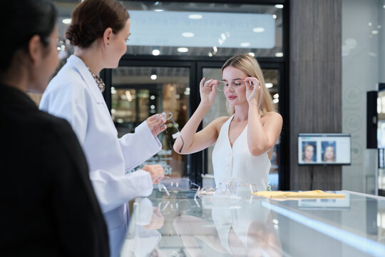White female customer chooses eyeglasses with optometrist's help and guidance, improved eyesight for individual lifestyles, consults on decisions, and options choices at store display in optical shop.