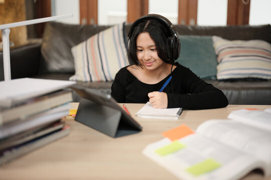 Asian teenage girl concentrates on online class, remote learning at home using tablet, writing and reading textbooks, self-study for back to school, Homeschooling, university education lifestyle.