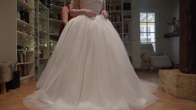 Bride in white wedding dress being helped in cozy indoor room with bookshelves