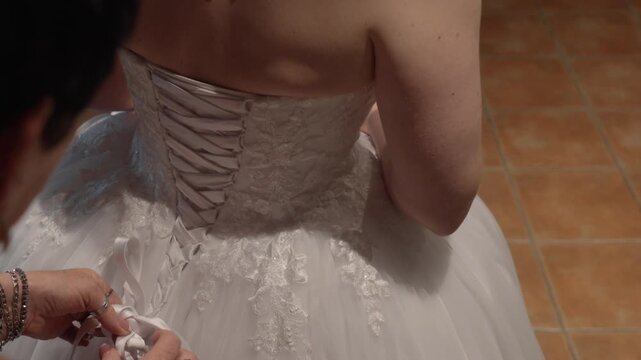 Hands tying the lace back of a white wedding dress on tiled indoor floor