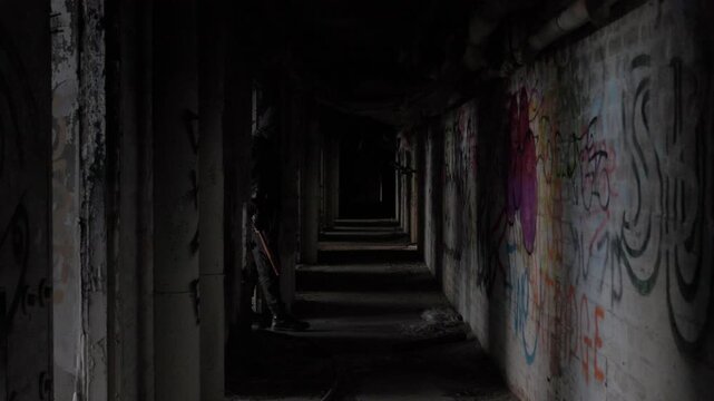 Suspicious shadowy figure carrying a wooden stick into and through dark abandoned factory graffiti-covered corridor, creating an atmospheric scene of mystery, fear and suspense