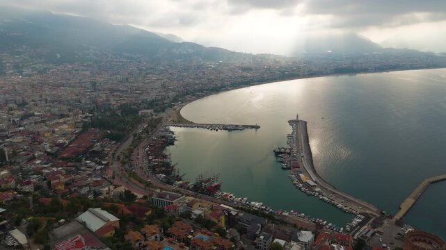 Alanya, Turkey. Aerial panoramic view of the historic harbor, Red Tower fortress, and Mediterranean coastline with dramatic sun rays piercing low clouds. Aerial View